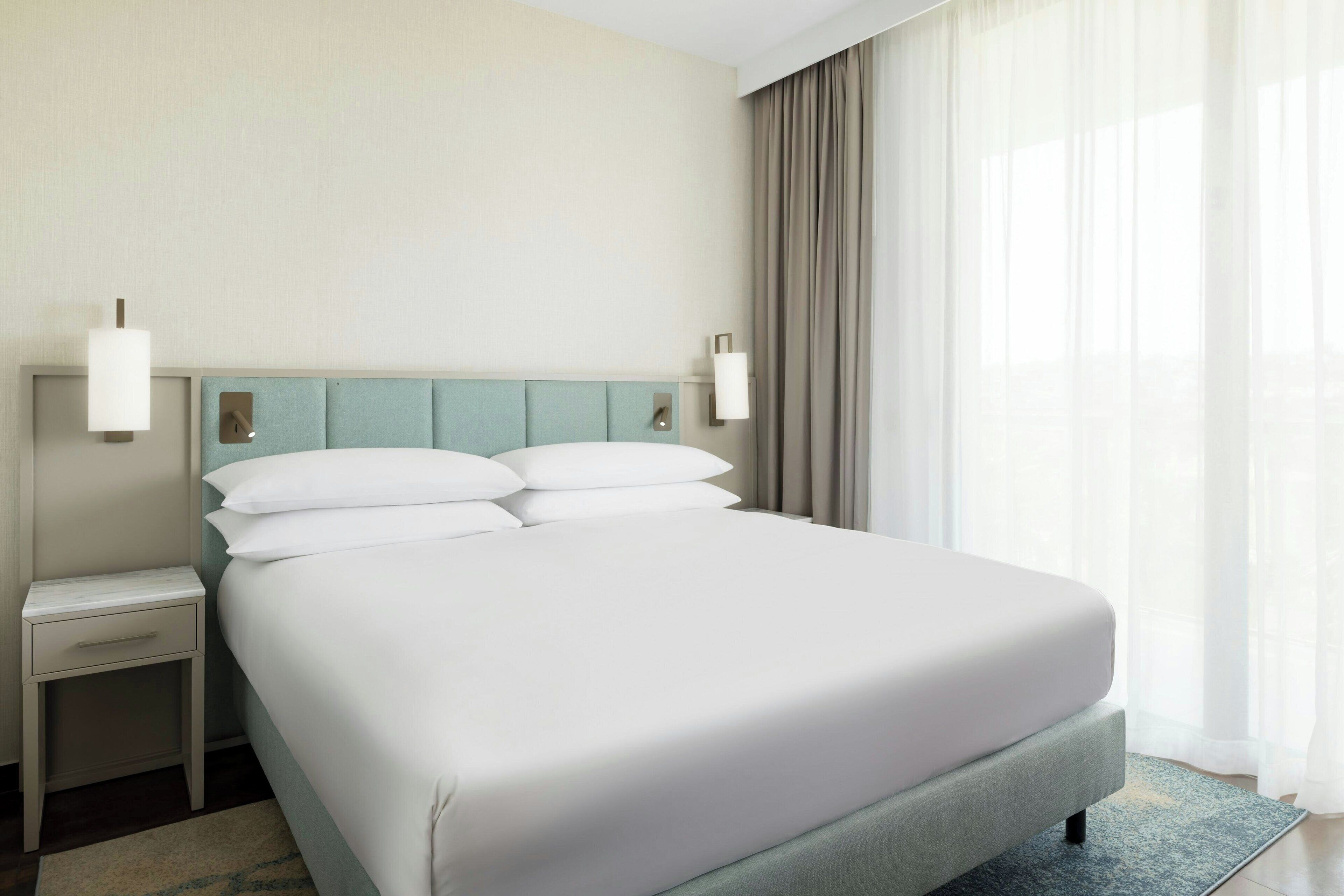 Een netjes opgemaakt bed met witte lakens, een lichtblauwe hoofdbord en een klein nachtkastje met lade.