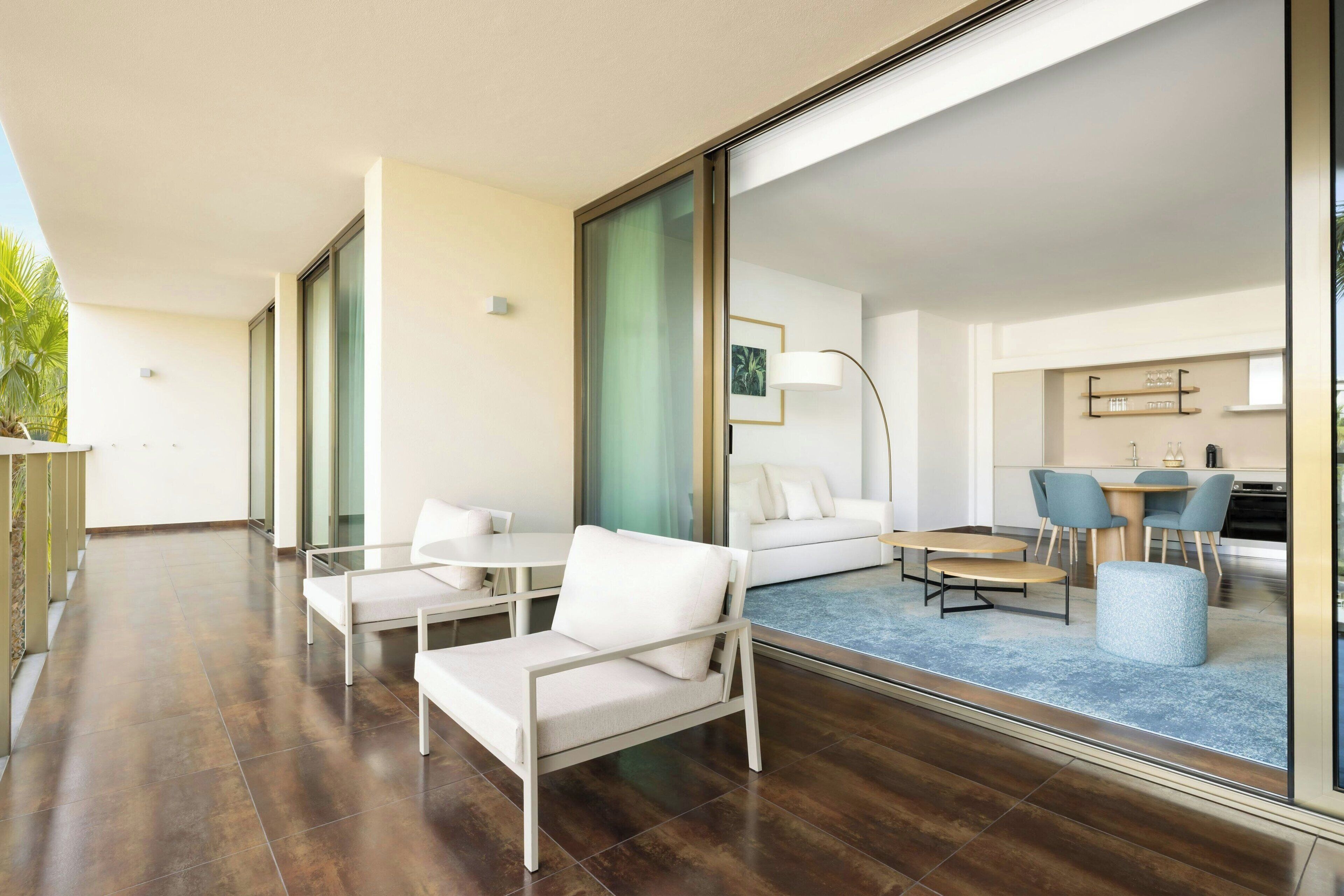 A modern hotel room with a large sliding glass door leading to a balcony, a white sofa, a small table, and a blue carpeted area.