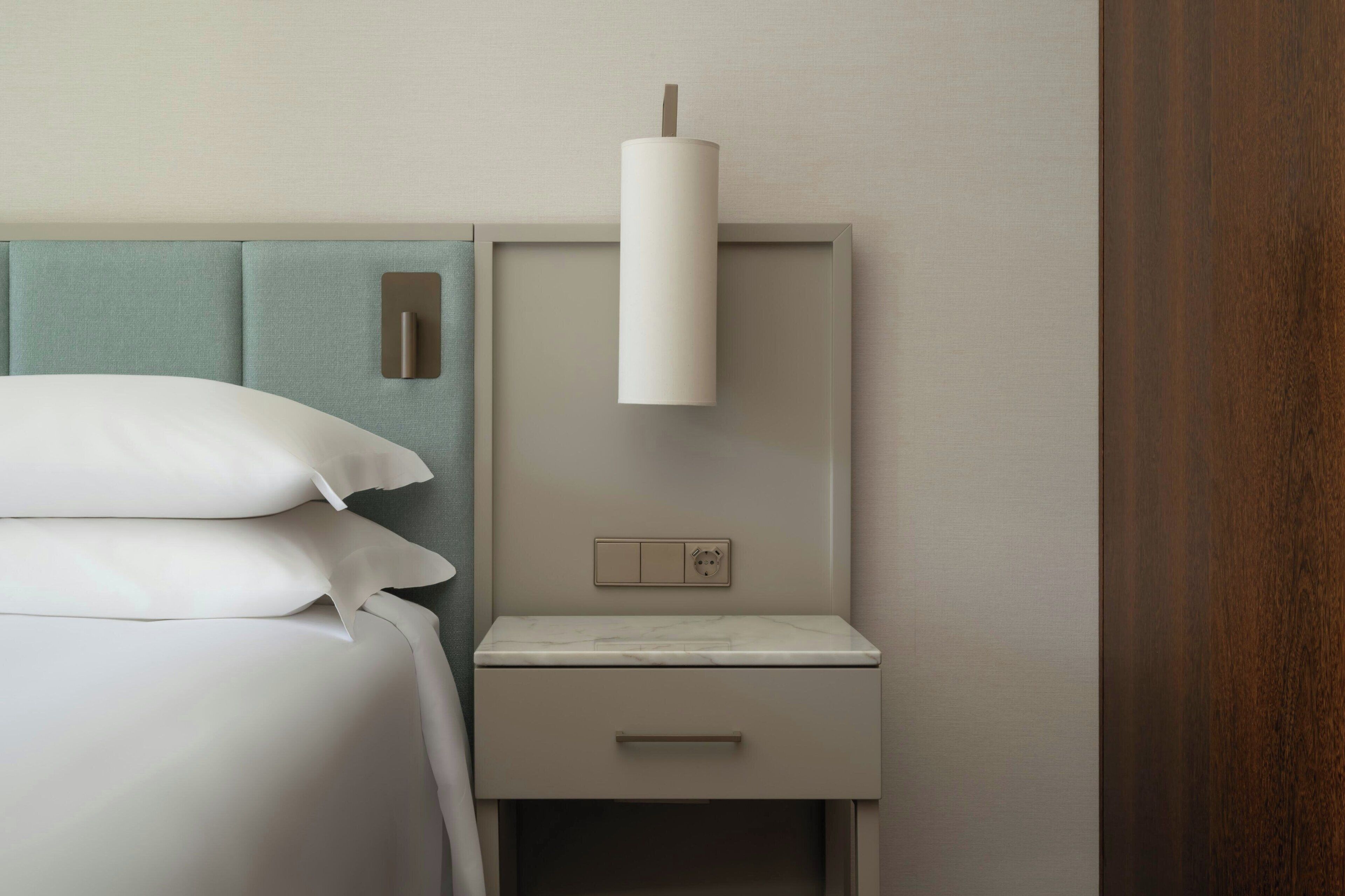 Un lit bien fait, avec du linge de lit blanc, une tête de lit, une table de chevet dotée d’un tiroir et un luminaire fixé au mur.