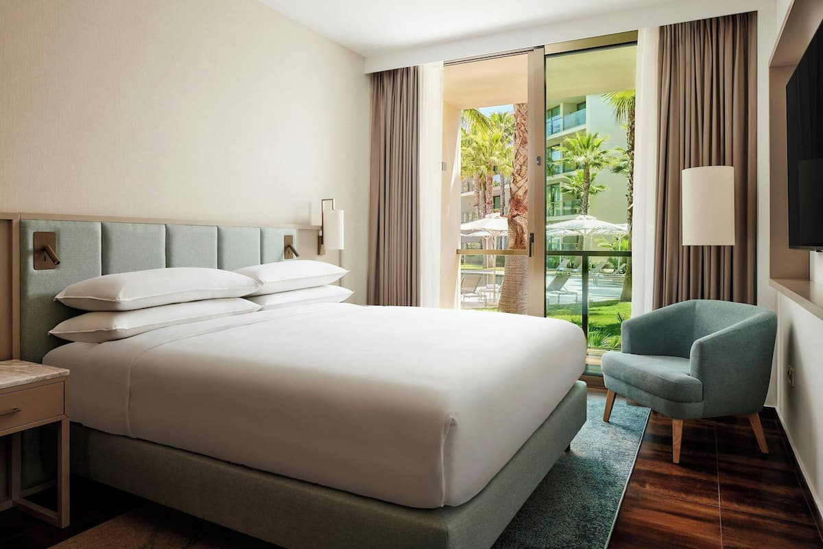 Ein modernes Hotelzimmer mit einem großen Bett, einer Sitzecke und Blick auf den Außenpool und Palmen.