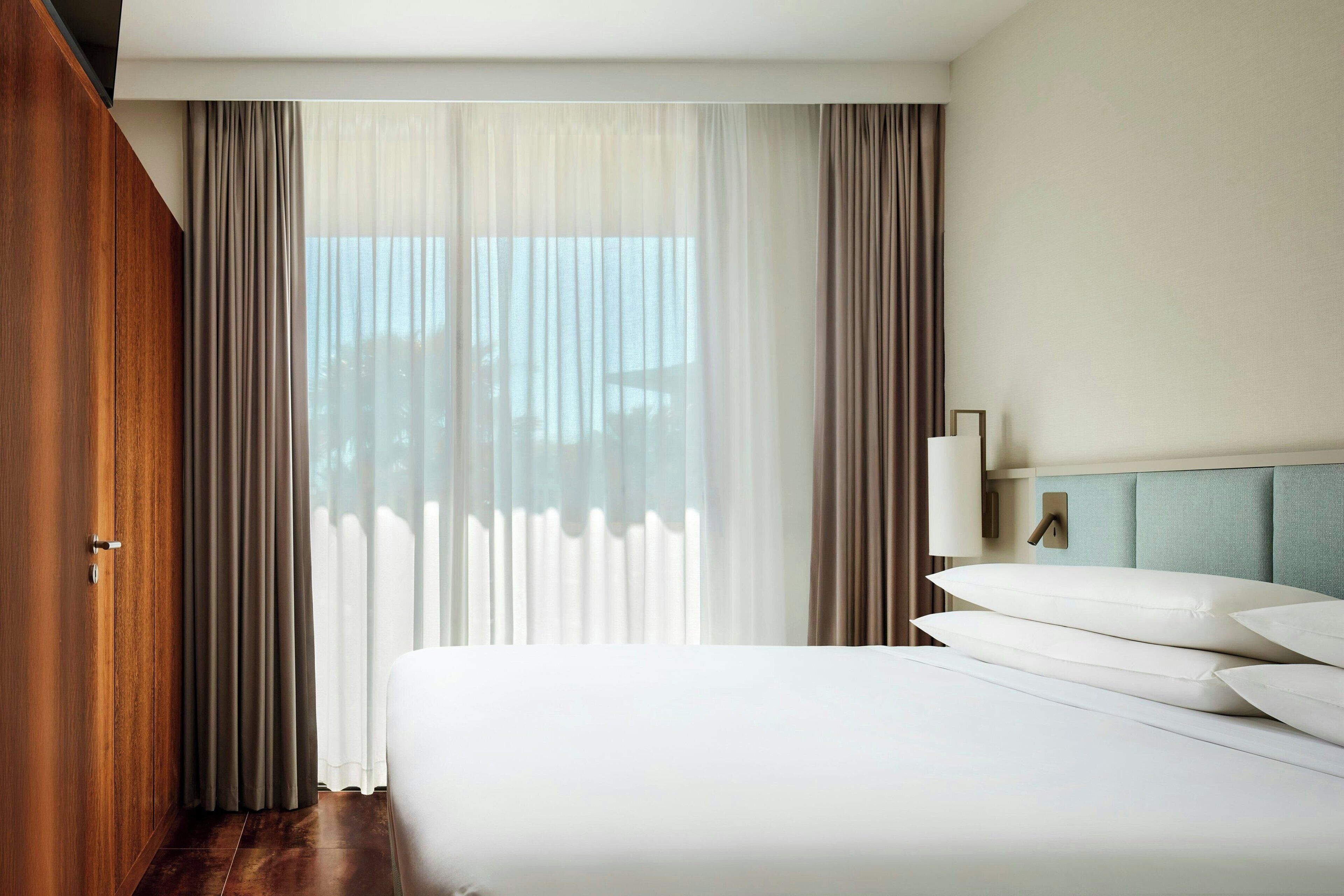 A hotel room with a large bed, a wooden wardrobe, a nightstand with a lamp, and sheer curtains.