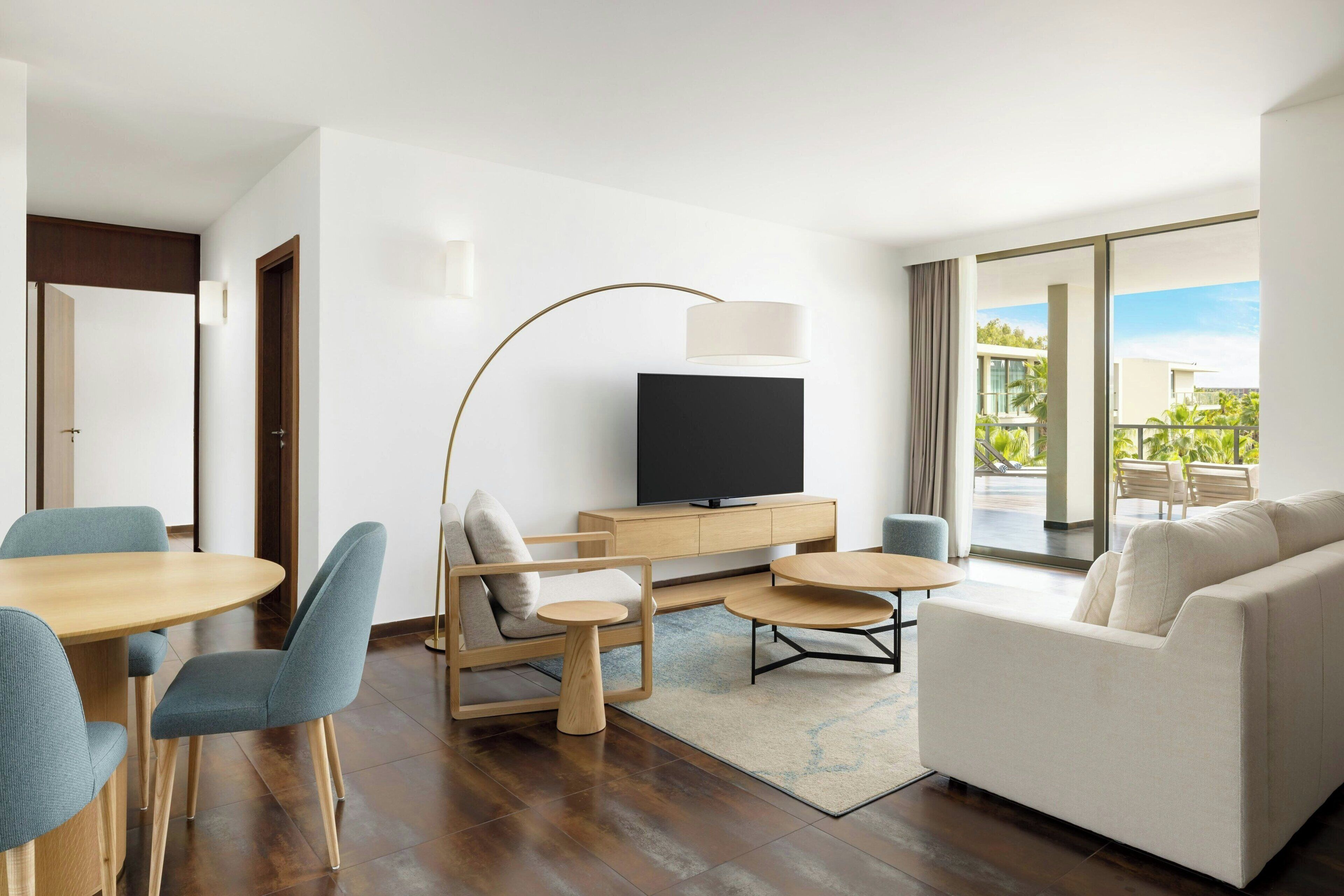 A modern hotel room with a flat-screen TV, a sofa, a dining table with chairs, and a large window offering a view of the outdoors.