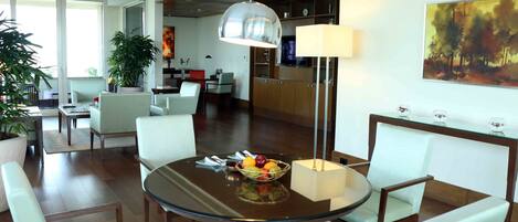 A modern dining area with a glass table, white chairs, a fruit bowl, and a pendant light.