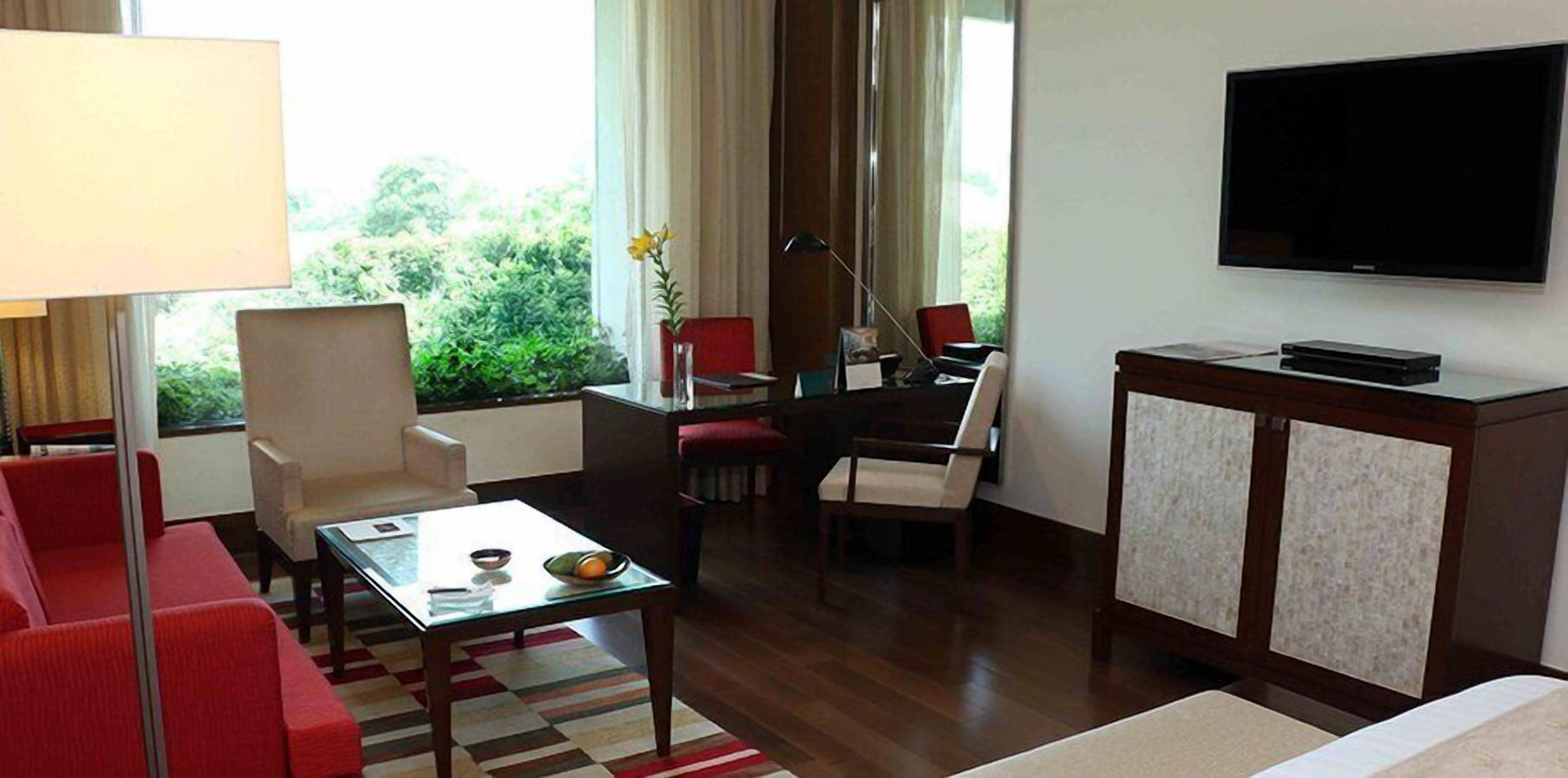 Una camera d'albergo con un divano rosso, un tavolino in vetro, un tavolo da pranzo con sedie, una TV a parete e una finestra con tende.