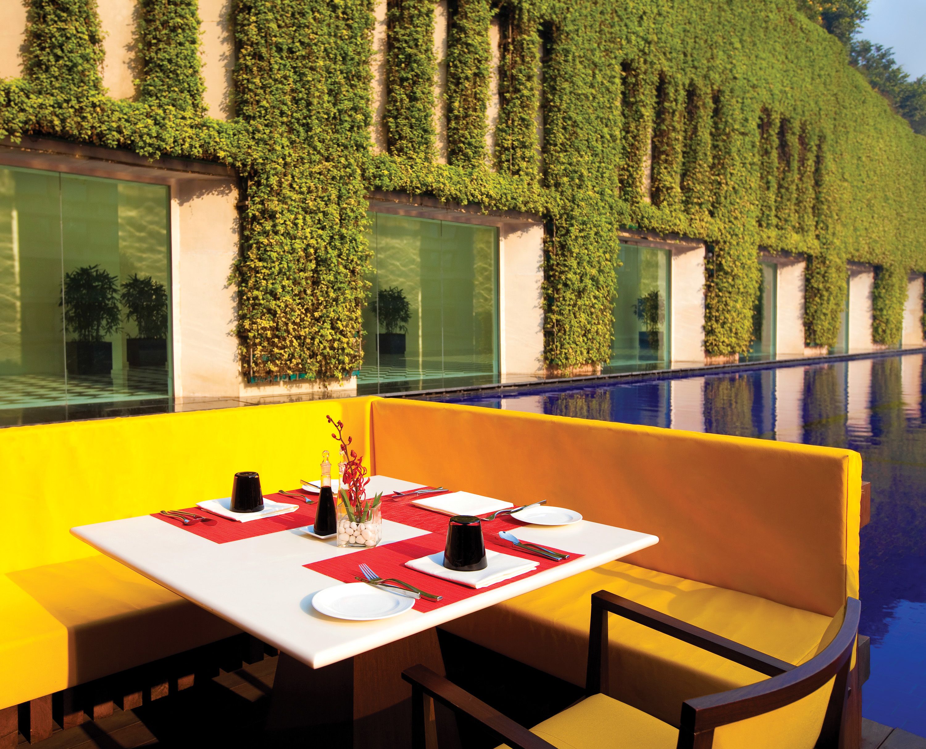 A dining area with a table set for two, a bench, and a view of a pool with a green wall in the background.