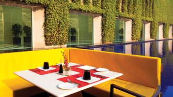 A dining area with a table set for two, a bench, and a view of a pool with a green wall in the background.