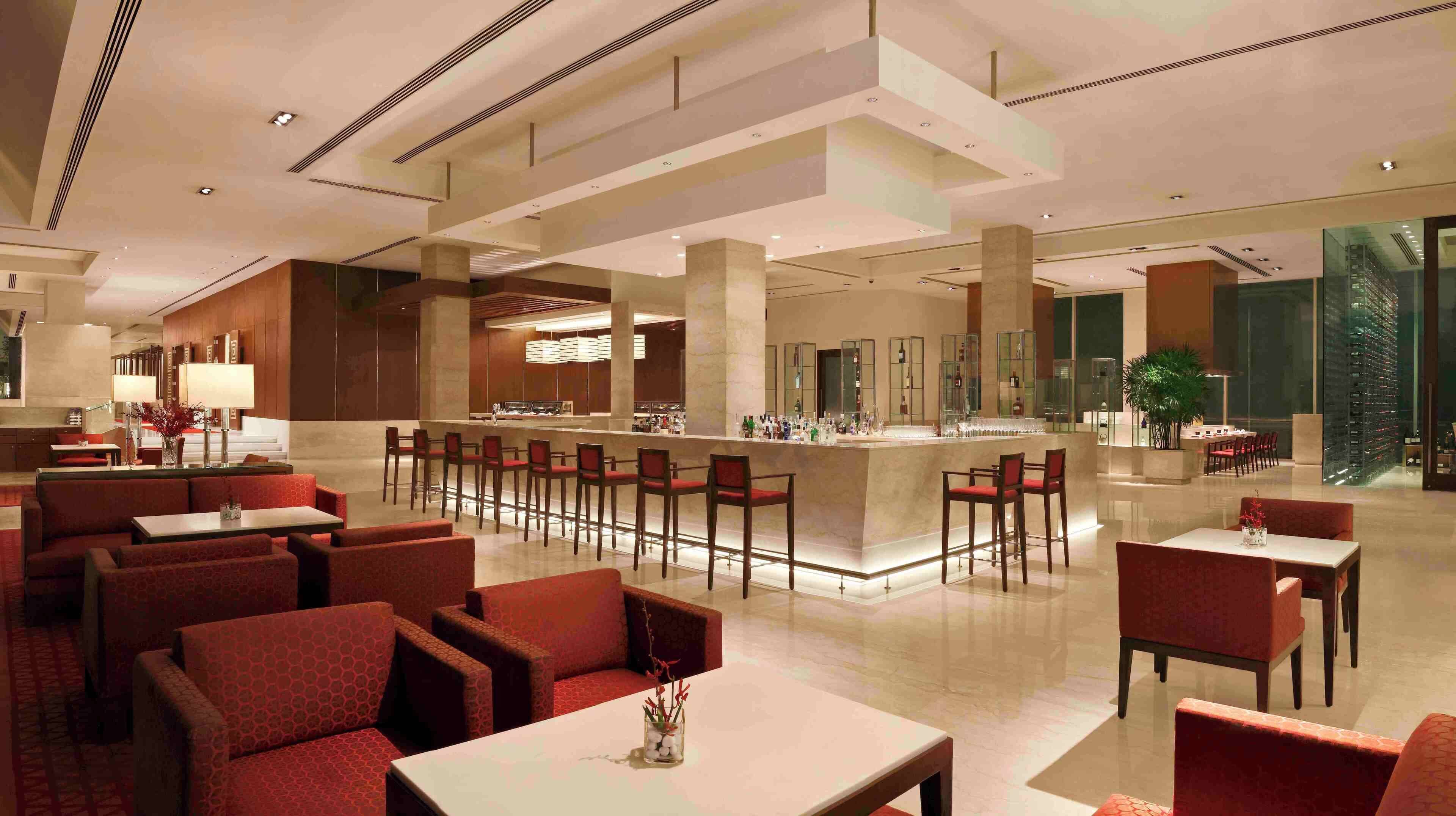 Un amplio vestíbulo de hotel con una zona de bar central, diversas opciones de asientos que incluyen cabinas y mesas, y una planta grande en la esquina.