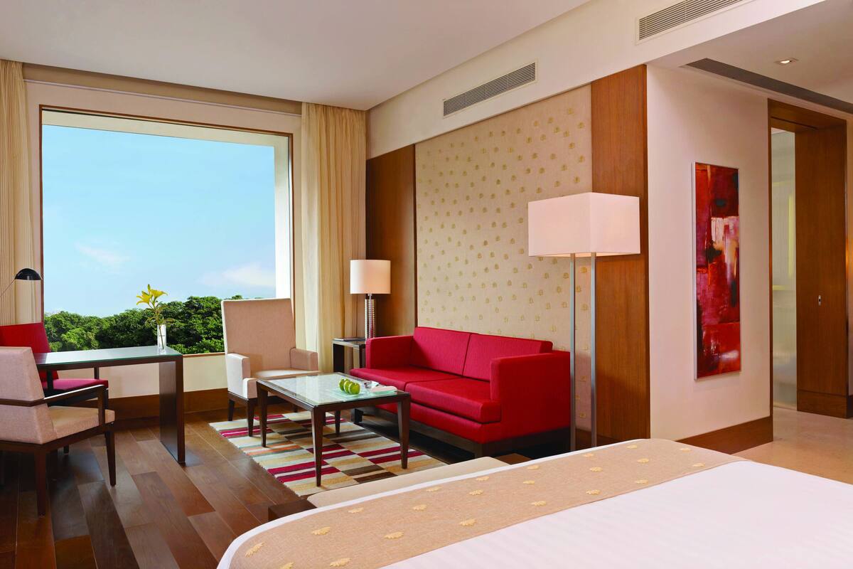 Quarto de hotel com uma cama, sofá vermelho, mesa pequena, cadeira, escrivaninha e uma grande janela com vista para o verde.