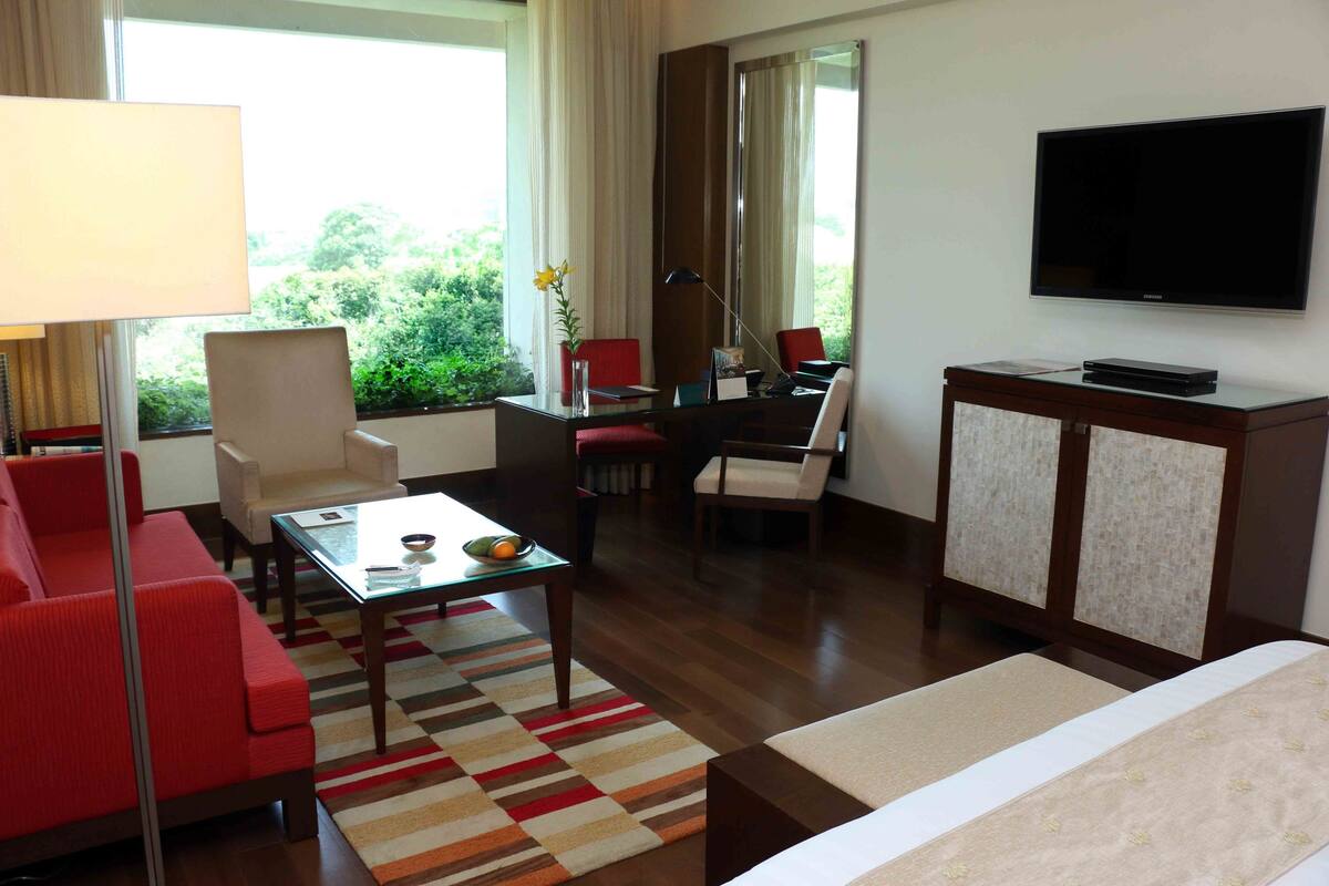 Quarto de hotel com sofá, poltronas, uma mesinha de centro com um vaso e frutas, uma televisão e um armário.