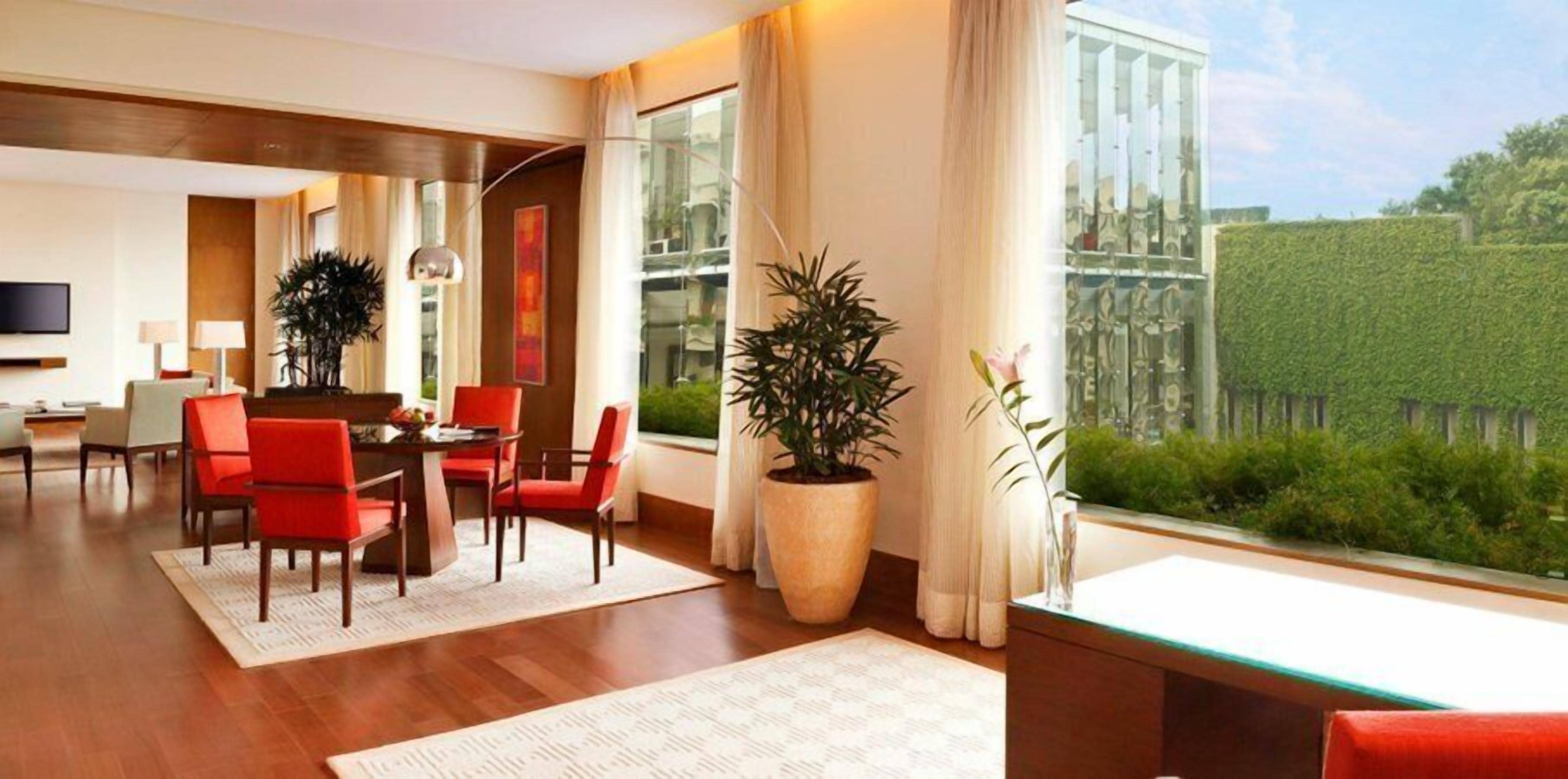 A modern hotel room with a dining area, large windows, and a view of the greenery outside.