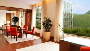 A modern hotel room with a dining area, large windows, and a view of the greenery outside.