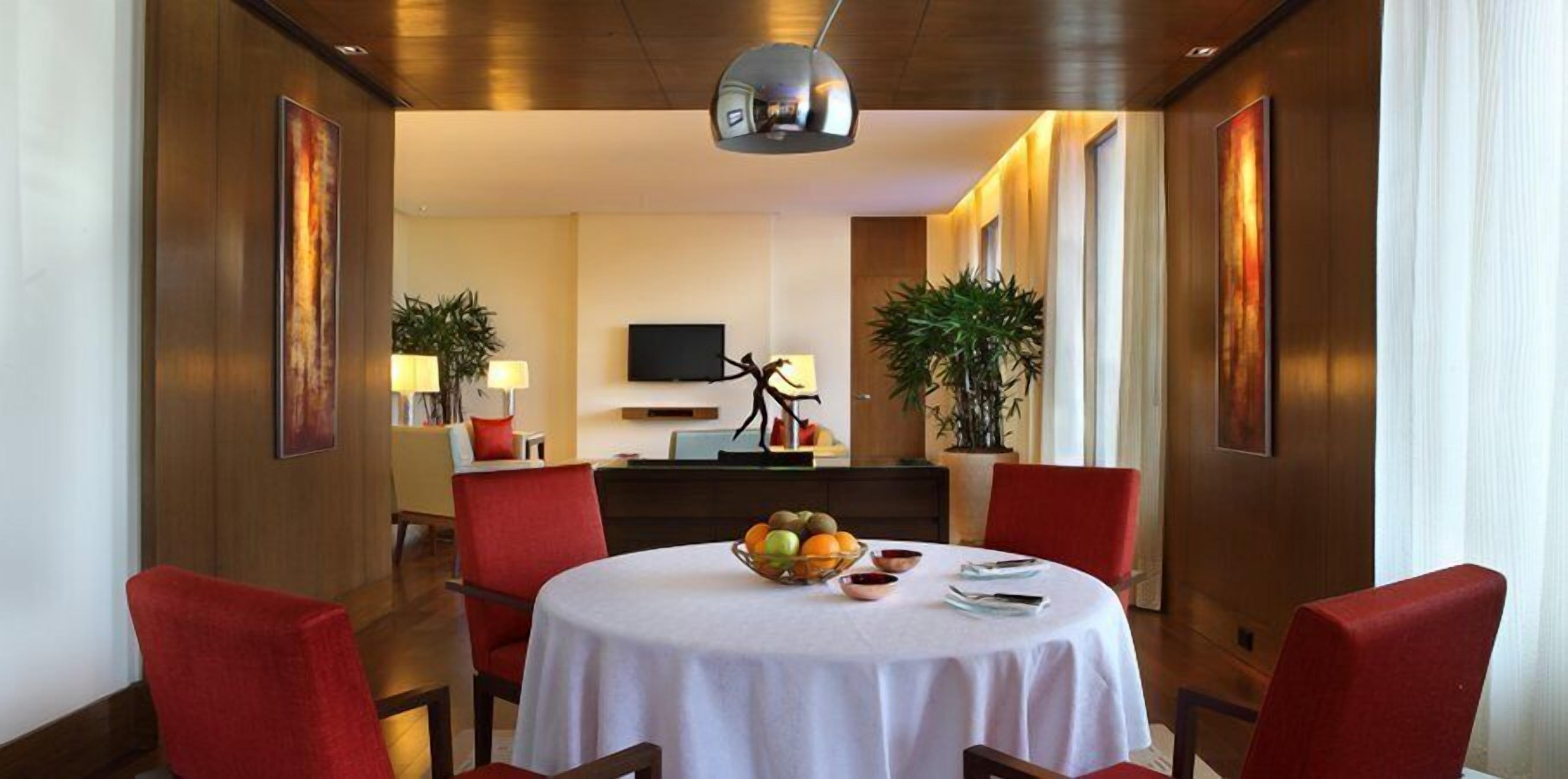 A dining area with a round table, red chairs, and a fruit bowl. There are two potted plants, a flat-screen TV on the wall, and a wooden ceiling.