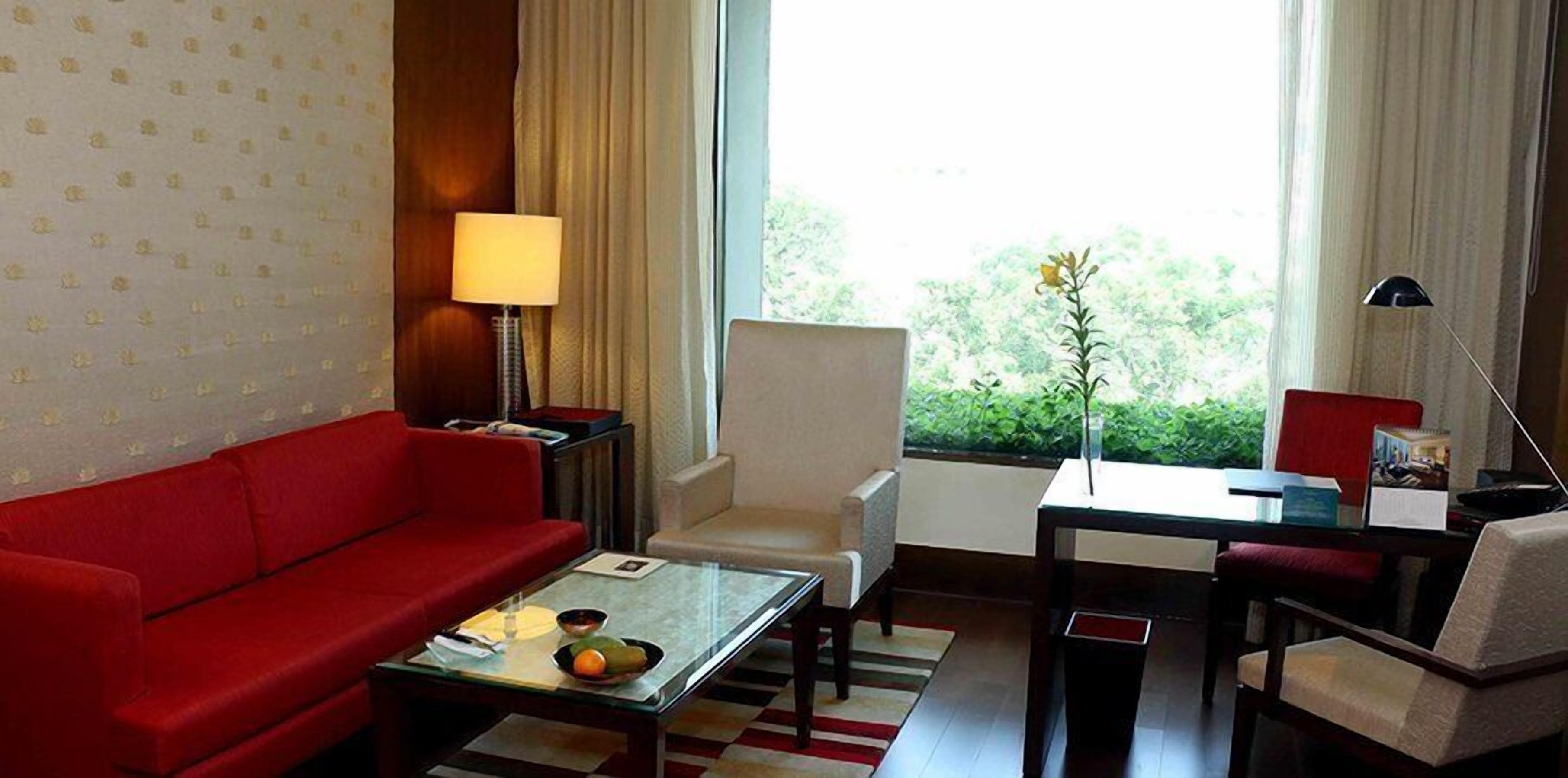 Habitación de hotel con sofá rojo, mesa de centro de vidrio, sillón blanco, pequeña mesa de comedor y vista a zonas verdes.