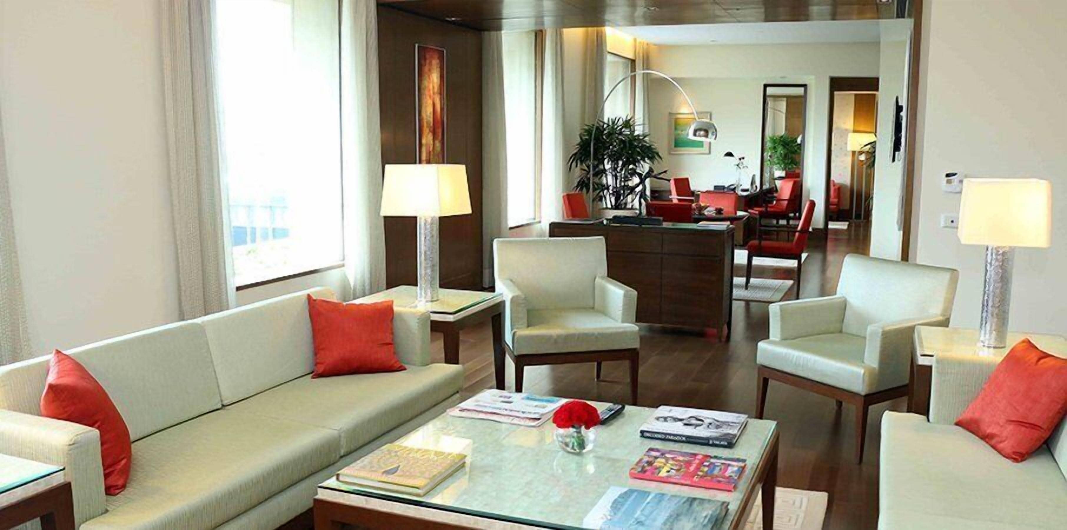 A modern hotel room with a sofa, armchairs, a coffee table, and a dining area in the background.