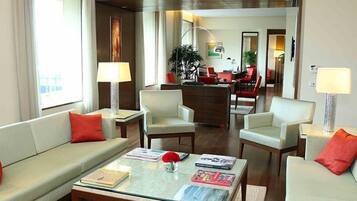 A modern hotel room with a sofa, armchairs, a coffee table, and a dining area in the background.