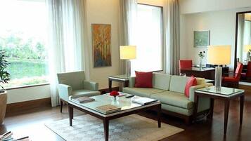 A well-lit living room with a sofa, armchairs, a coffee table, and a dining table. There is a large window with curtains, a painting on the wall, and a potted plant.
