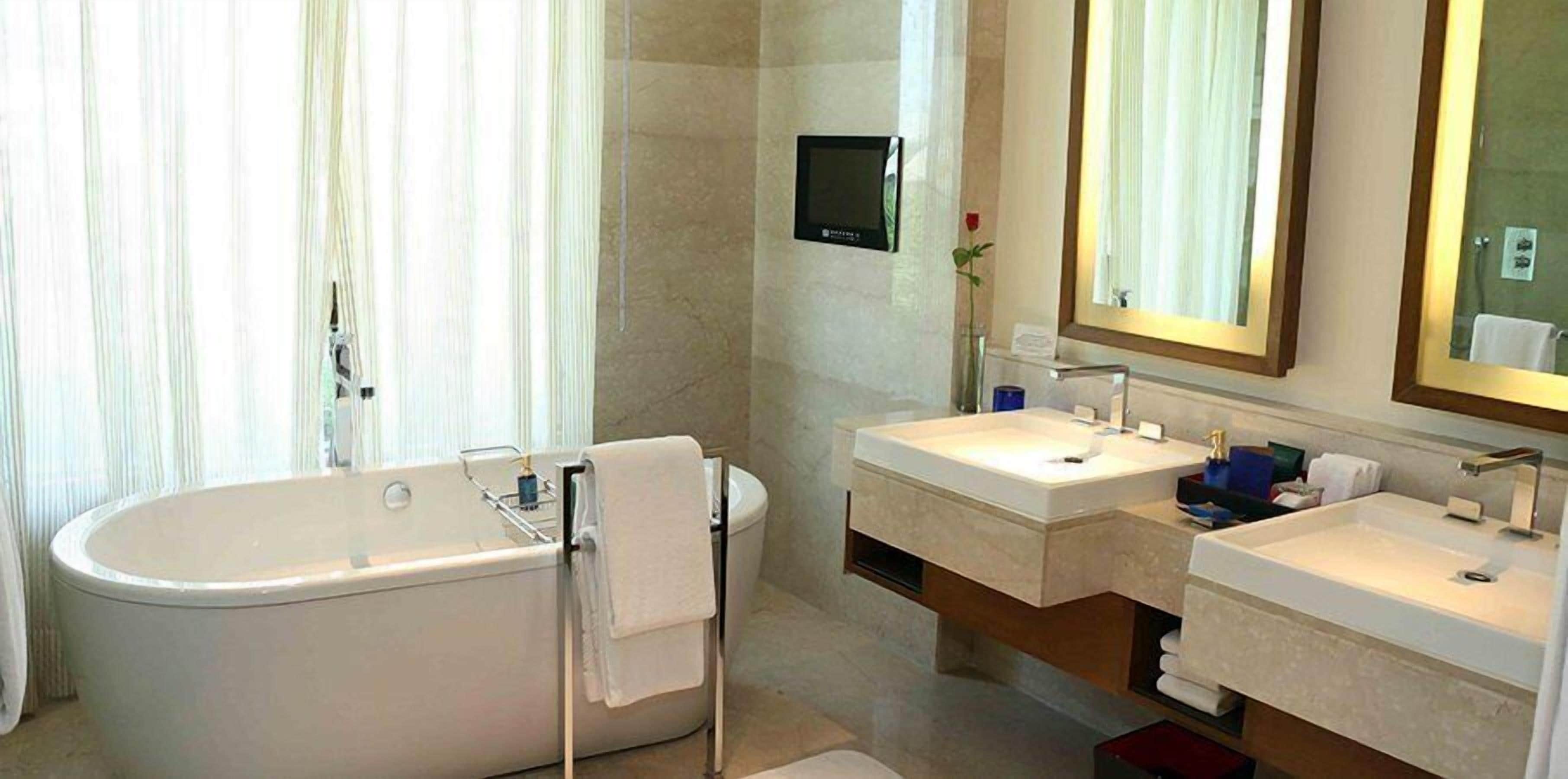 A modern bathroom with a freestanding bathtub, two sinks, a large mirror, and a television mounted on the wall.