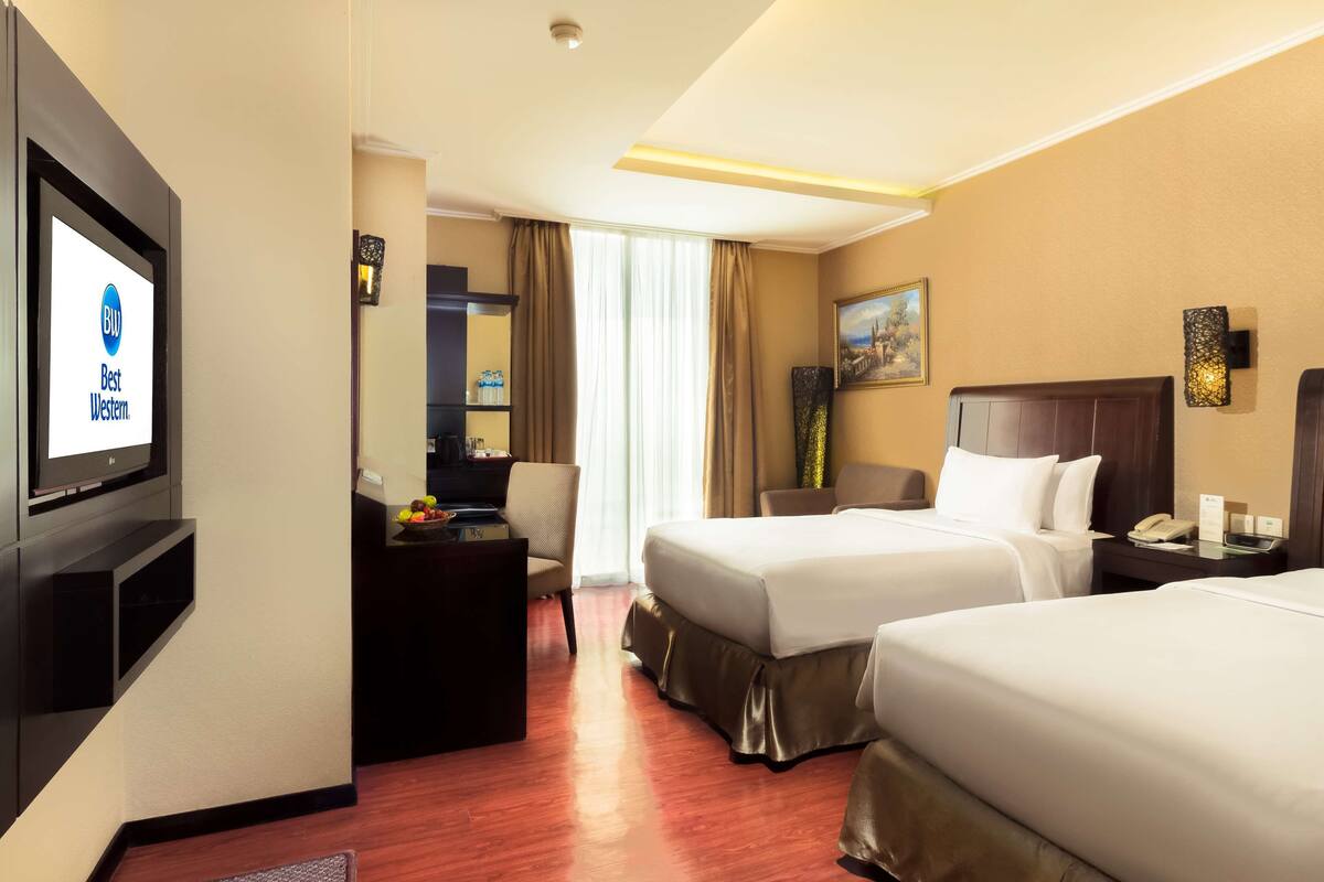 A hotel room with two beds, a desk, a TV, and a bathroom visible through a glass door.