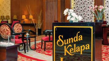 Eine Lobby mit rotem Teppich, einem Empfangstresen mit der Aufschrift 'Sunda Kelapa Lounge' und traditioneller Einrichtung, darunter Holzwände und gestaltete Tapeten.