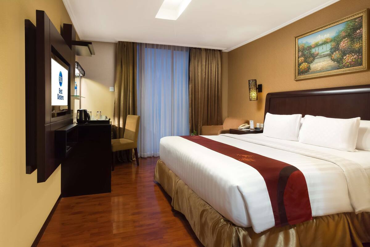 A hotel room with a large bed, a desk with a computer, a chair, a TV, and a painting on the wall.