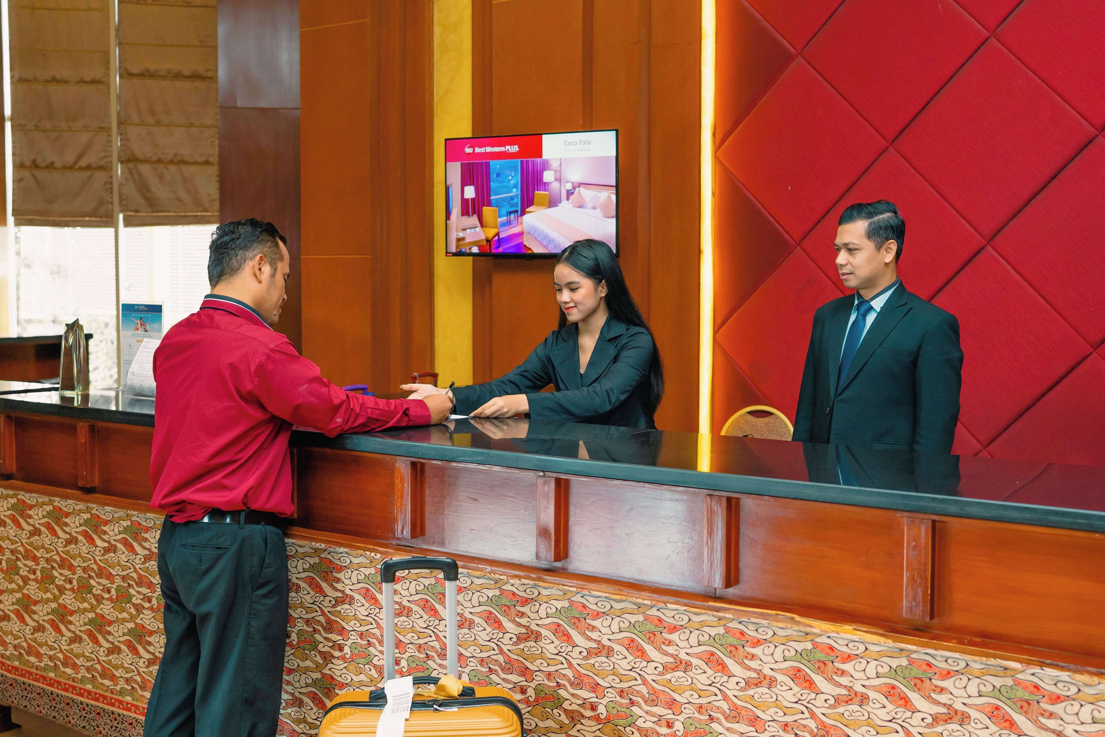 Un mostrador de recepción con una pantalla digital mostrando opciones de habitaciones, una alfombra estampada y una maleta en el suelo.