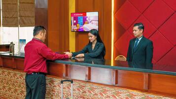 Un mostrador de recepción con una pantalla digital mostrando opciones de habitaciones, una alfombra estampada y una maleta en el suelo.