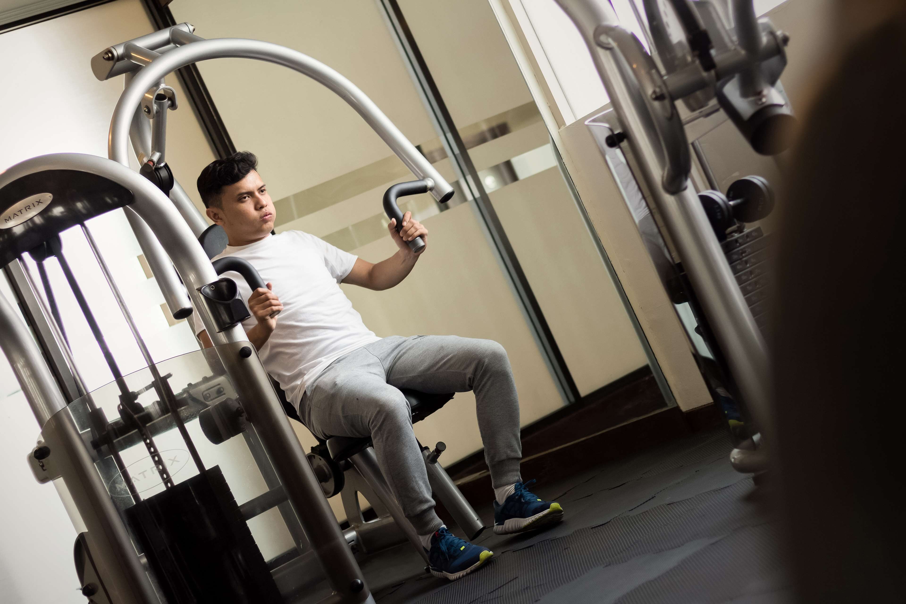 Um homem usando um aparelho de leg press numa academia com paredes espelhadas e uma pilha de pesos visível.