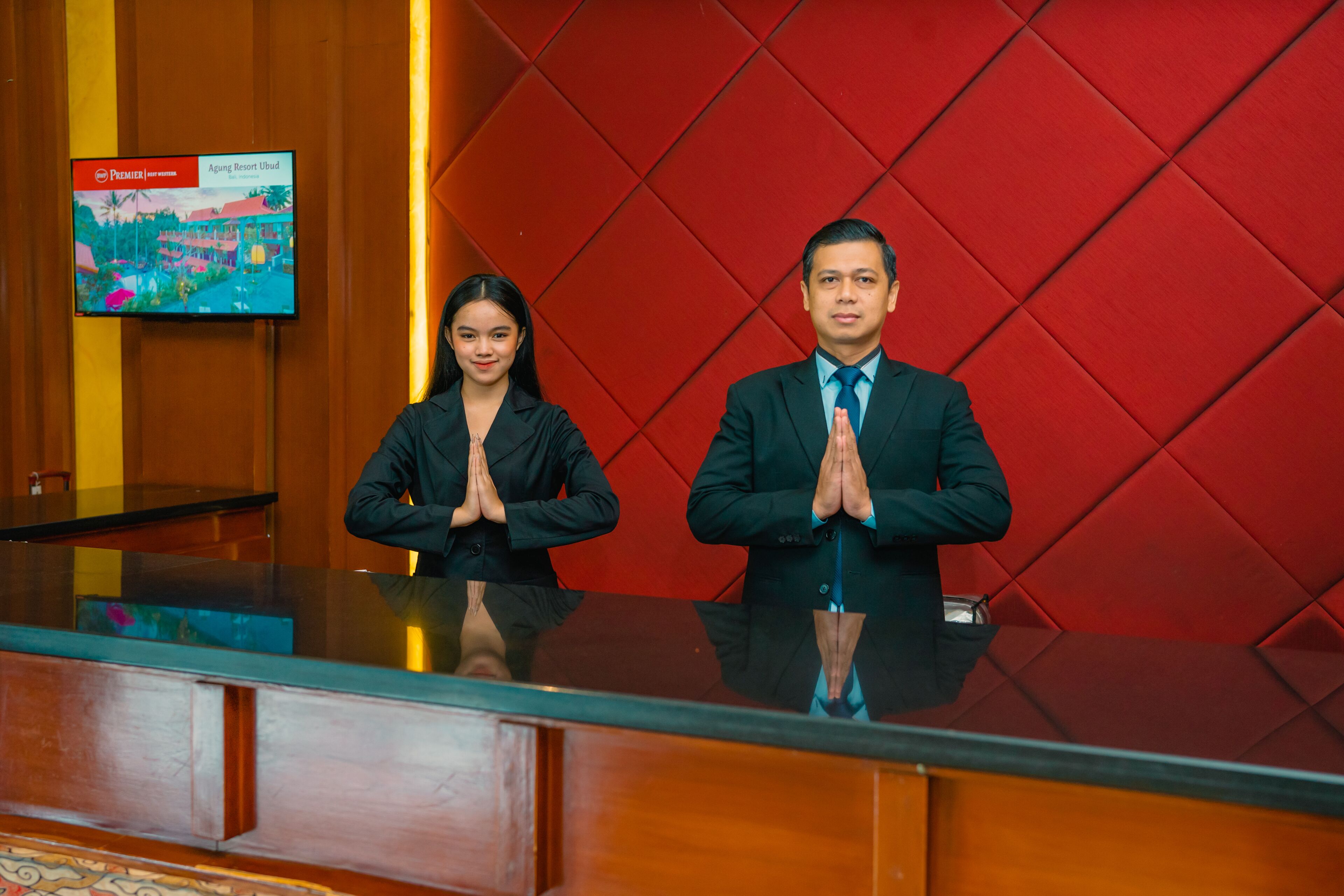 Two individuals standing at a reception desk with a red padded wall and a television displaying a resort image.