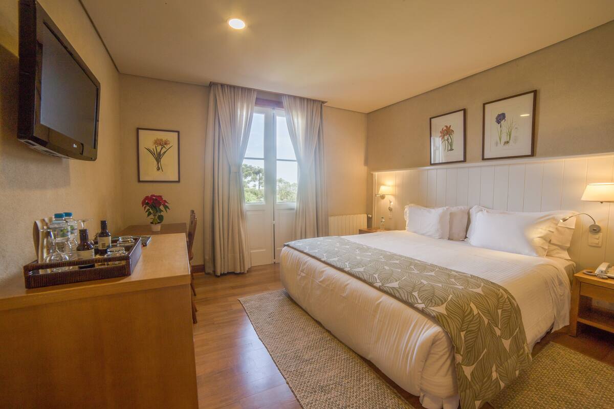 Une chambre d’hôtel avec un grand lit, un bureau sur lequel se trouve un plateau avec des articles de toilette, une télévision et, au mur, des œuvres d’art florales encadrées.