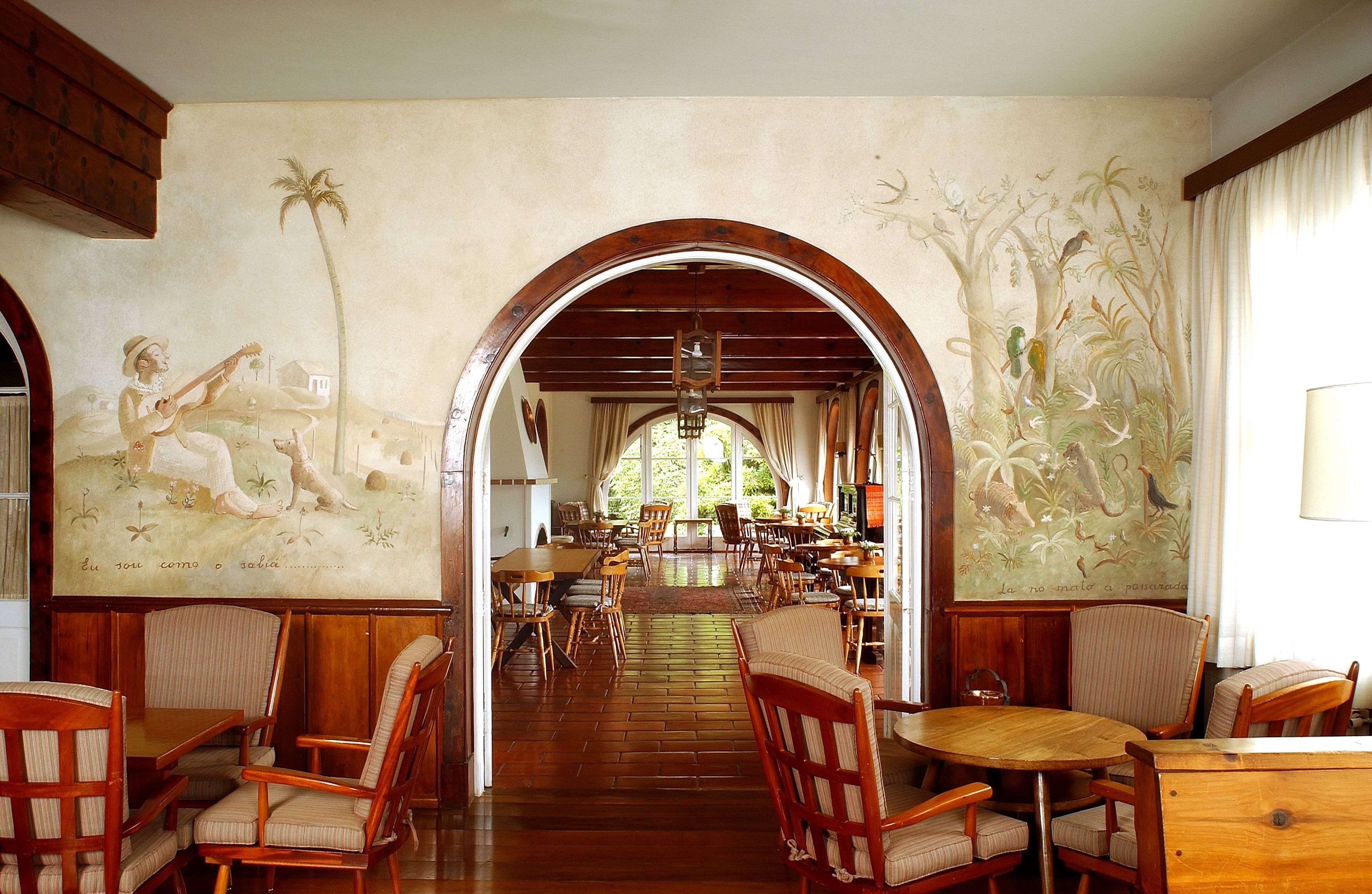 Un comedor amplio con mesas y sillas de madera, un techo alto y murales en las paredes que representan escenas tropicales.