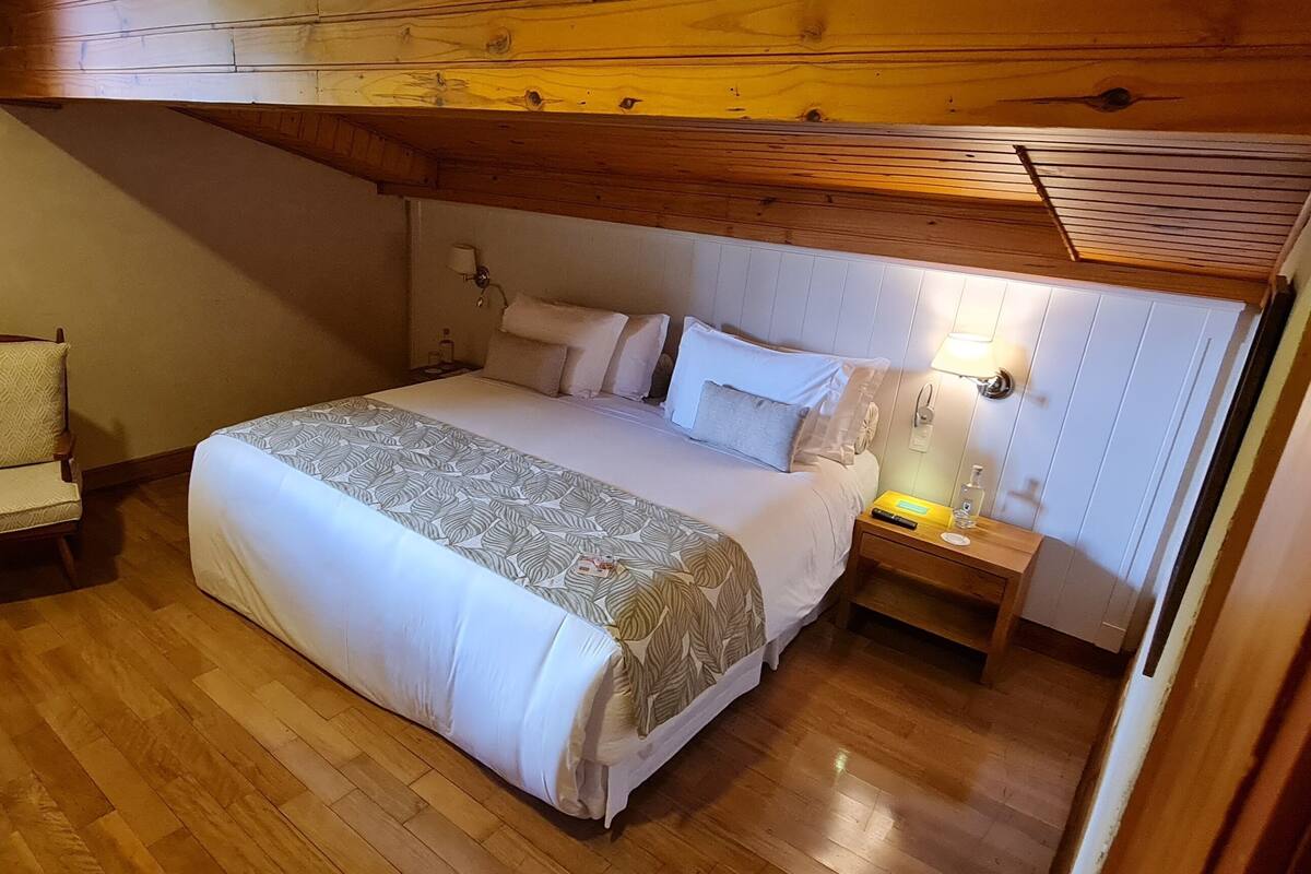 Une chambre avec un plafond en bois, un lit recouvert d’une couvre-lit à motifs, une table de chevet avec une lampe et une chaise.