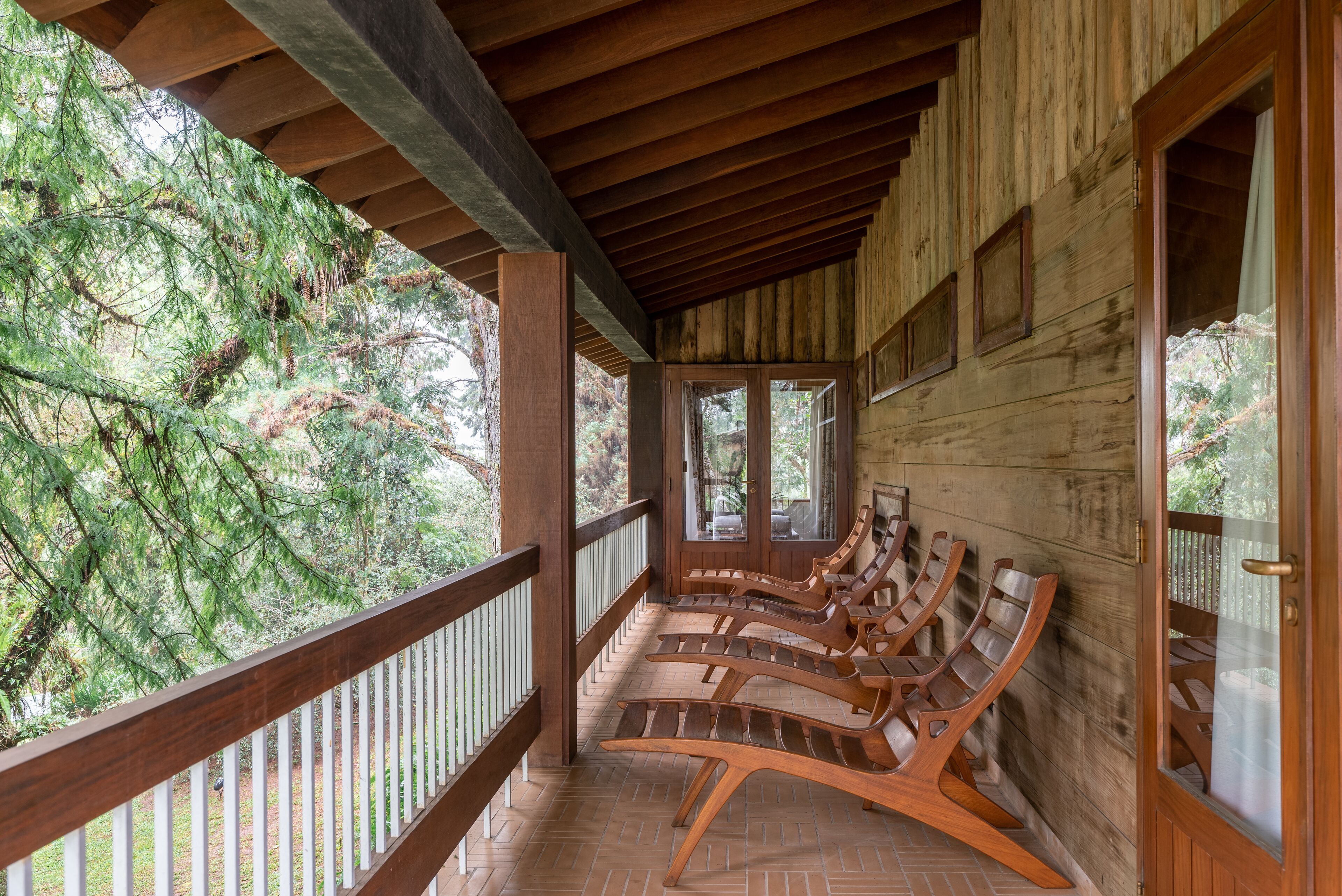 A wooden porch with a railing, wooden chairs, and a view of trees.