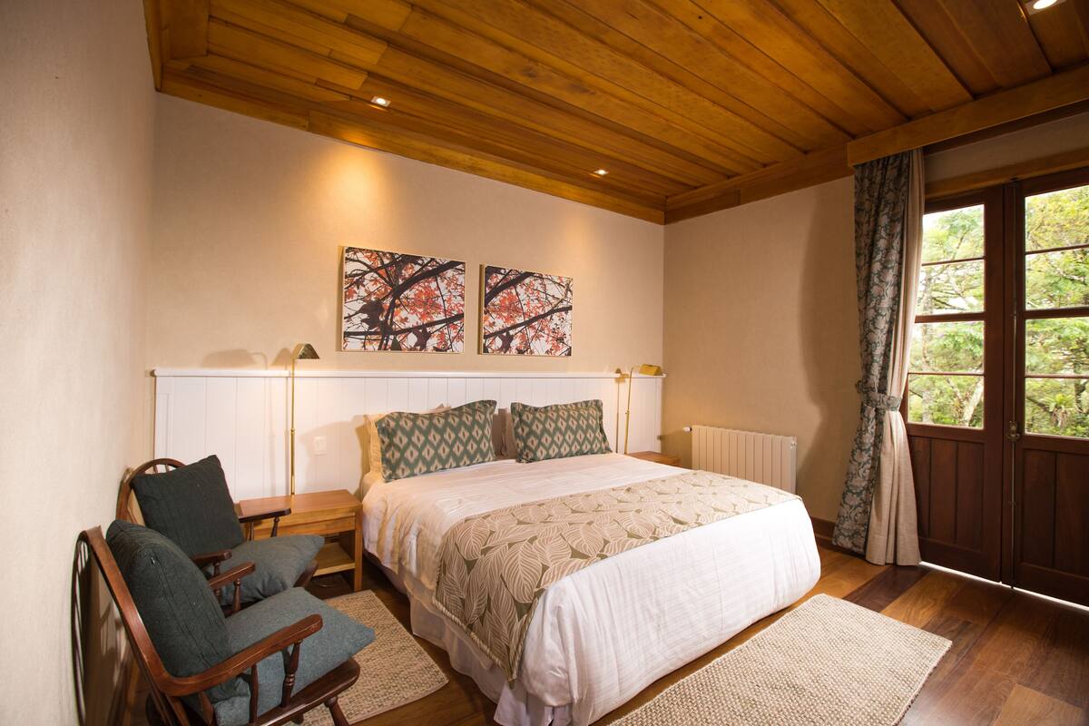 Une chambre avec un plafond en bois, un grand lit avec une literie à motifs, deux tableaux accrochés au mur, une chaise en bois et une fenêtre avec des rideaux.