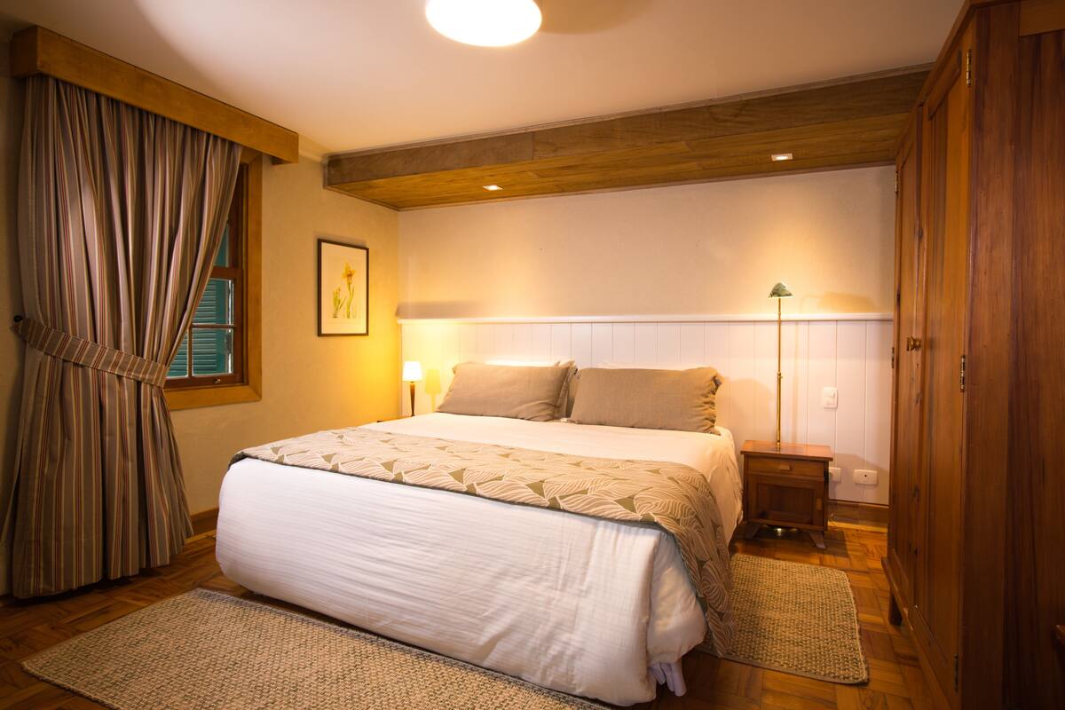 Une chambre à coucher avec un grand lit, une tête de lit en bois, une table de chevet, une lampe sur pied et une fenêtre avec des rideaux.