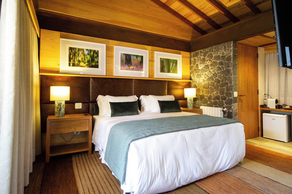 Une chambre bien éclairée avec un plafond en bois, un mur d’accent en pierre, un lit avec une literie blanche, deux tables de chevet avec des lampes et trois tableaux encadrés au-dessus du lit.