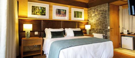Un dormitorio bien iluminado con techo de madera, una pared de acento de piedra, una cama con ropa blanca, dos mesitas de noche con lámparas y tres cuadros enmarcados sobre la cama.