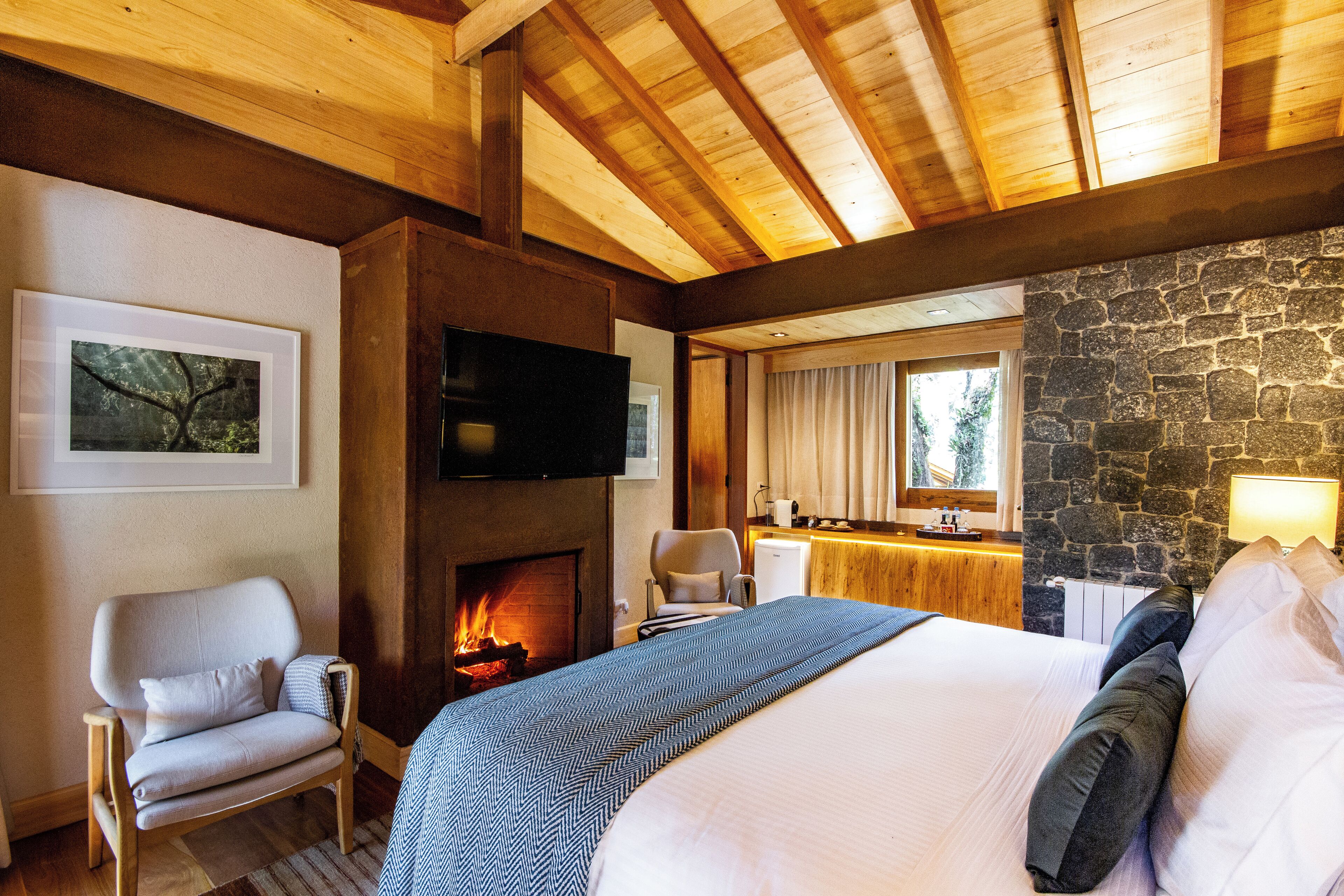 Quarto de hotel com uma cama grande, lareira, televisão, parede de pedra e teto de madeira.
