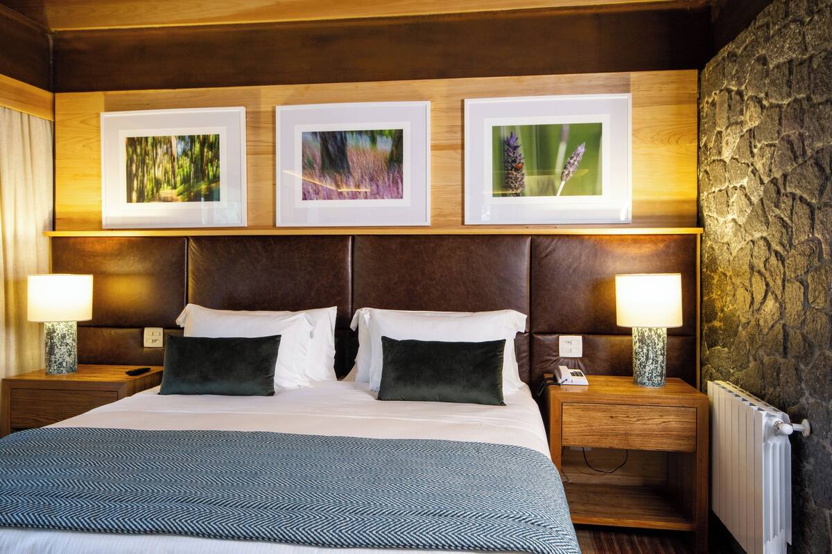 Une chambre d’hôtel avec un grand lit, une tête de lit en bois, trois tableaux encadrés au mur, deux tables de chevet avec des lampes et un mur en pierre.