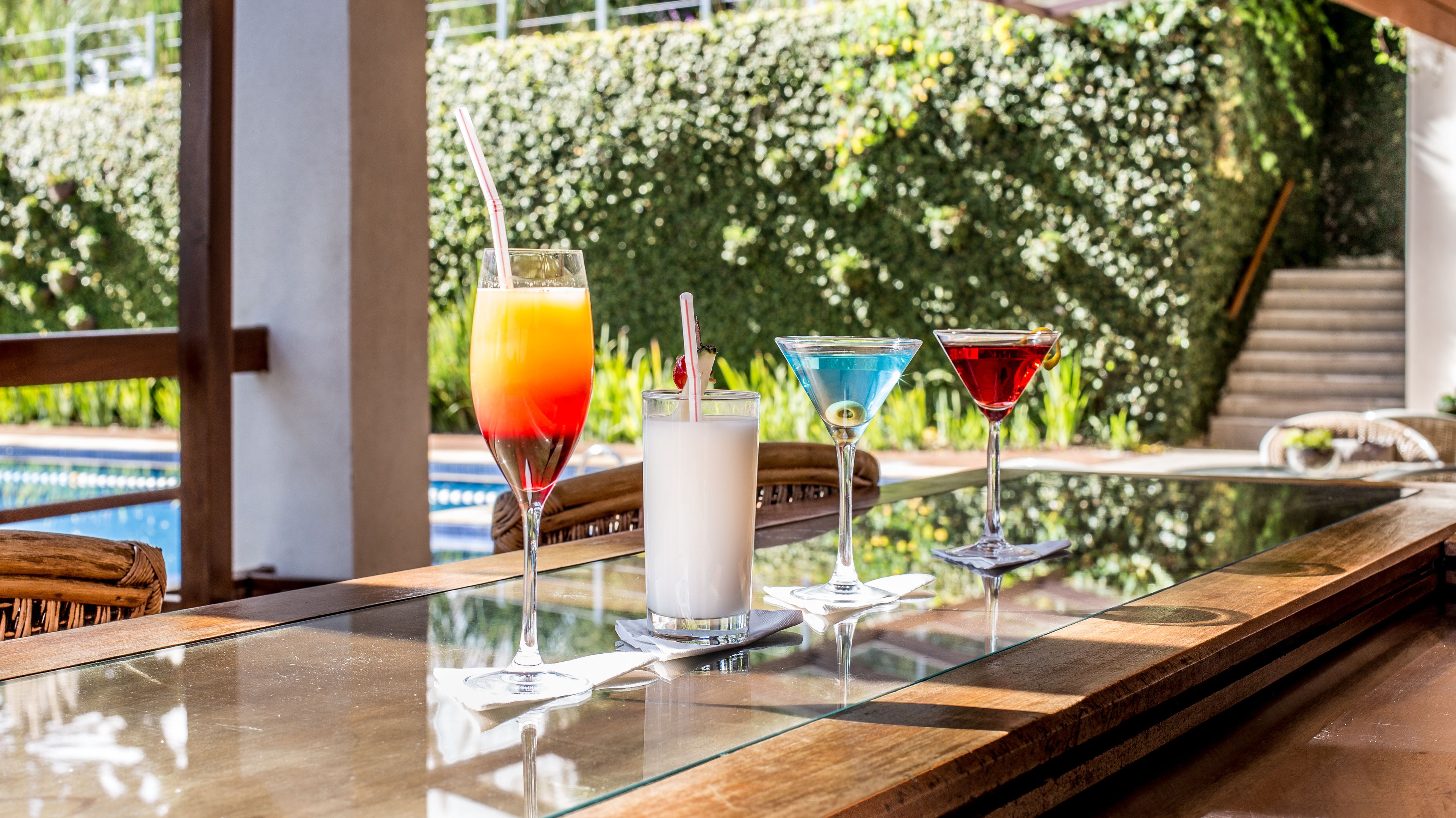 Zona de bar con tres bebidas sobre una mesa de cristal, piscina al fondo y terraza de madera.