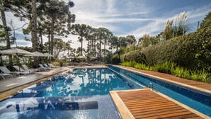 Una piscina con sillas de descanso y sombrillas, rodeada de árboles altos y una terraza de madera.