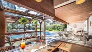 Zona de bar moderna con una mesa de vidrio, tres bebidas y vistas a una piscina y asientos exteriores.