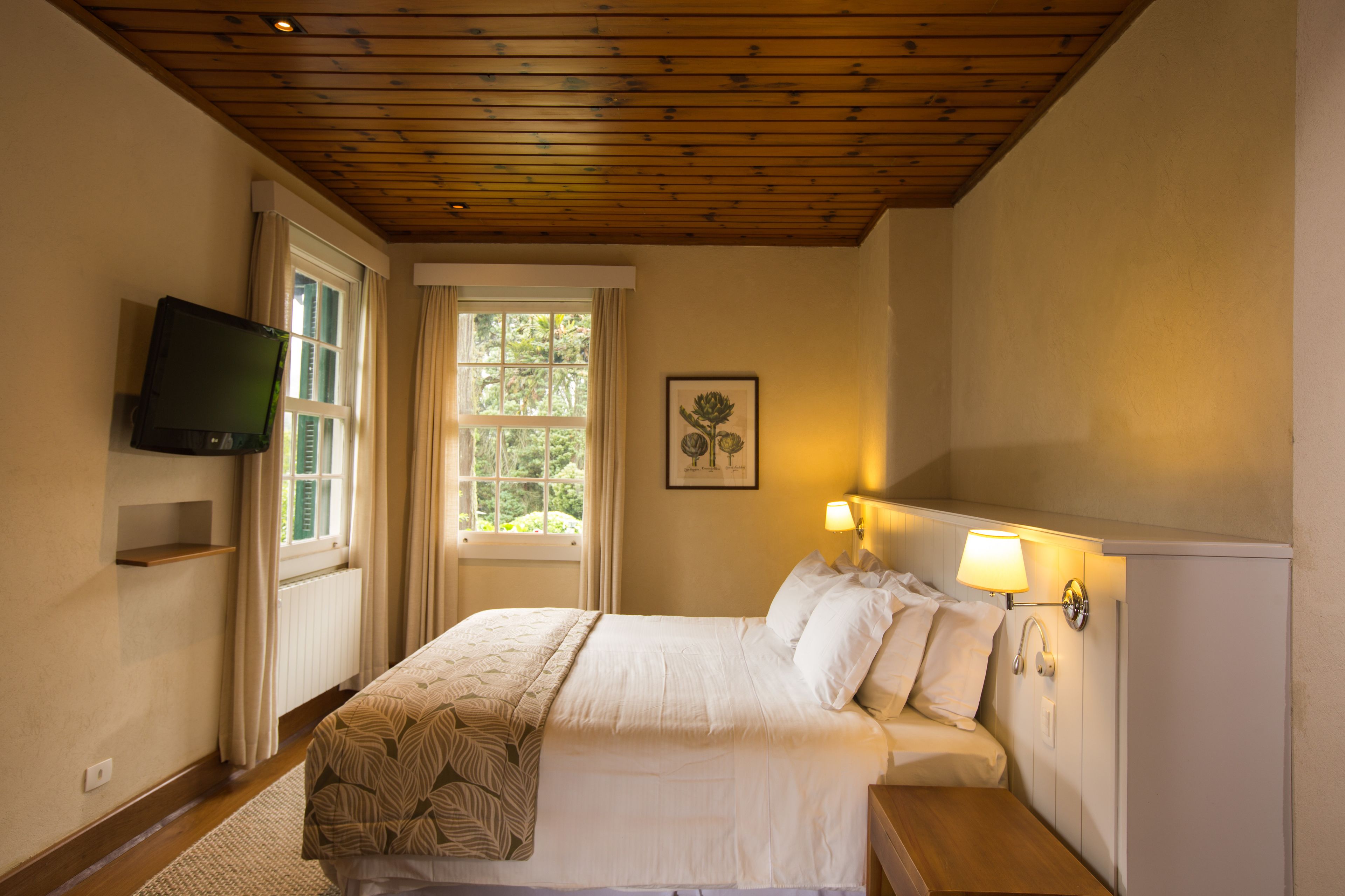 A bedroom with a wooden ceiling, a bed with white bedding, a bedside table with a lamp, a TV mounted on the wall, and a window with curtains.