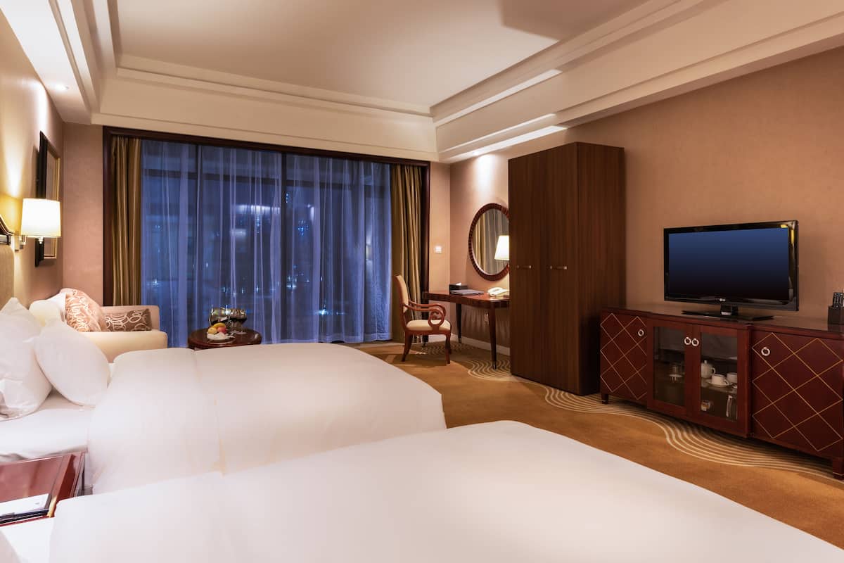 A hotel room with two beds, a television, a desk with a chair, a mirror, and a view of the city at night.