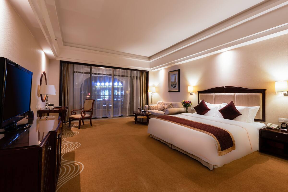 A hotel room with a large bed, a TV, a desk with a lamp, and a view of the city through the window.