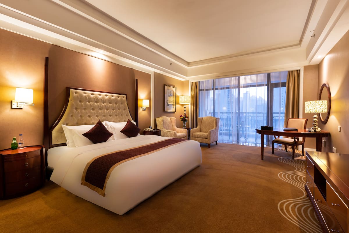 A hotel room with a large bed, a desk, a chair, a TV, and a view of the city through large windows.