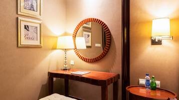 Een hotelkamer met een houten bureau, een ronde spiegel, een wandlamp, een klein rond tafeltje met flessen en afbeeldingen in lijsten aan de muur.