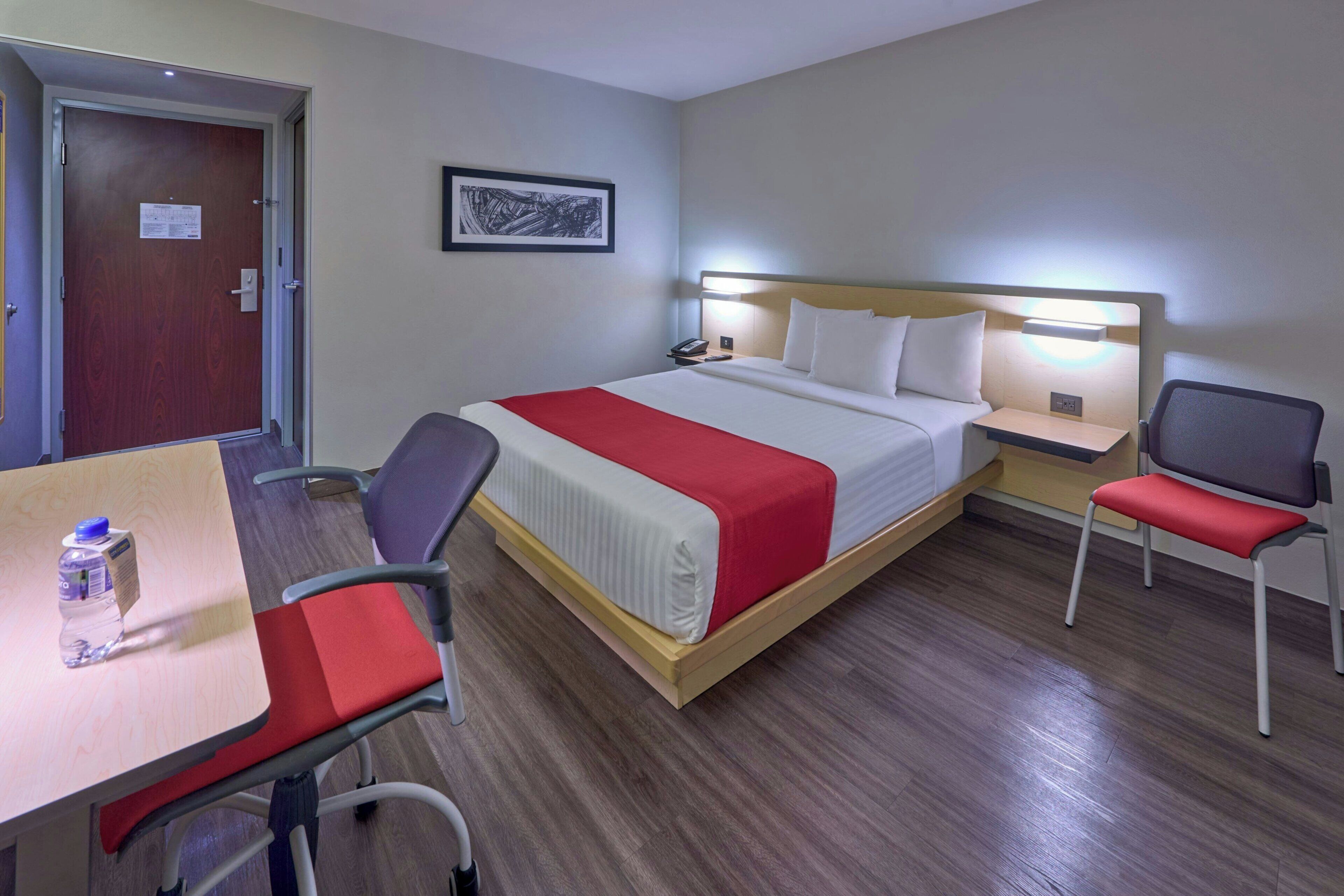 A hotel room with a bed, bedside table, chair, desk, and a water bottle on the desk.