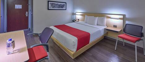 A hotel room with a bed, bedside table, chair, desk, and a water bottle on the desk.