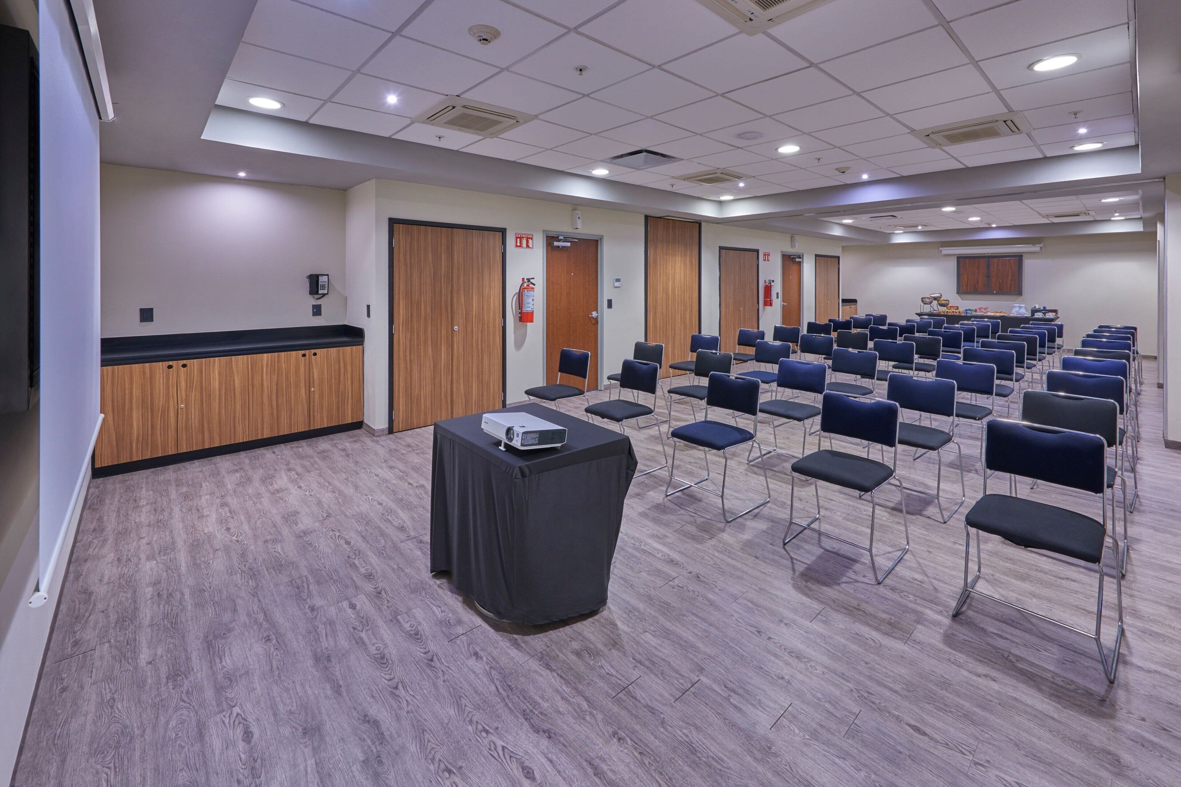 A conference room with rows of chairs, a table with a projector, wooden paneling, and fire safety equipment.