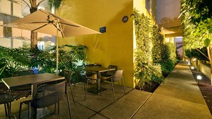 An outdoor patio area with a table and chairs, surrounded by greenery and a yellow wall. - City Express by Marriott San Luis Potosi Zona Universitaria (San Luis Potosi)